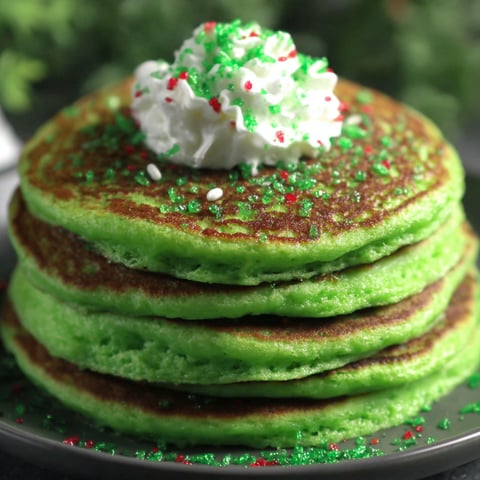 A stack of green pancakes with white whipped cream and red sprinkles.